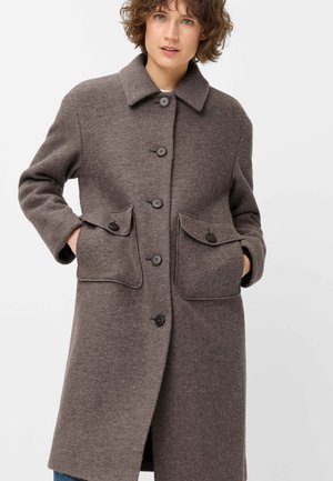 Woman wearing a long brown wool coat with large front buttoned pockets and black buttons, standing with hands in pockets.