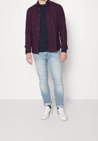 Camicia a quadri rossa e blu sopra un maglione blu navy, abbinata a jeans azzurri chiari e sneaker bianche con dettagli neri.