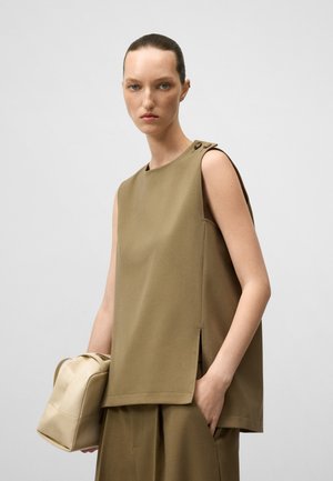 Donna con capelli tirati all'indietro, che indossa un top senza maniche e pantaloni verde oliva abbinati, mentre tiene una borsa in pelle beige su uno sfondo neutro.