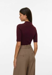 Mujer con cabello largo y oscuro, vistiendo una blusa ajustada de manga corta color burdeos y pantalones de pierna ancha beige de cintura alta, mirando de lado.