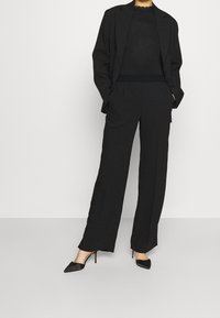 Svart skräddarsydd kostym med en löst sittande kavaj och högmidjade byxor. Ovandelen har ett texturerat band vid midjan. Svarta spetsiga skor.