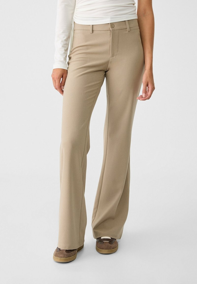 Mujer con pantalones beige de pierna recta, zapatos marrones sin cordones y una camiseta blanca de manga larga, de pie contra un fondo liso.