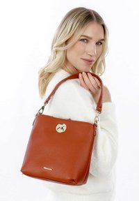 Bolso de piel color óxido con acabado texturizado, herrajes dorados y una correa desmontable. Presenta un cierre circular en la parte frontal.