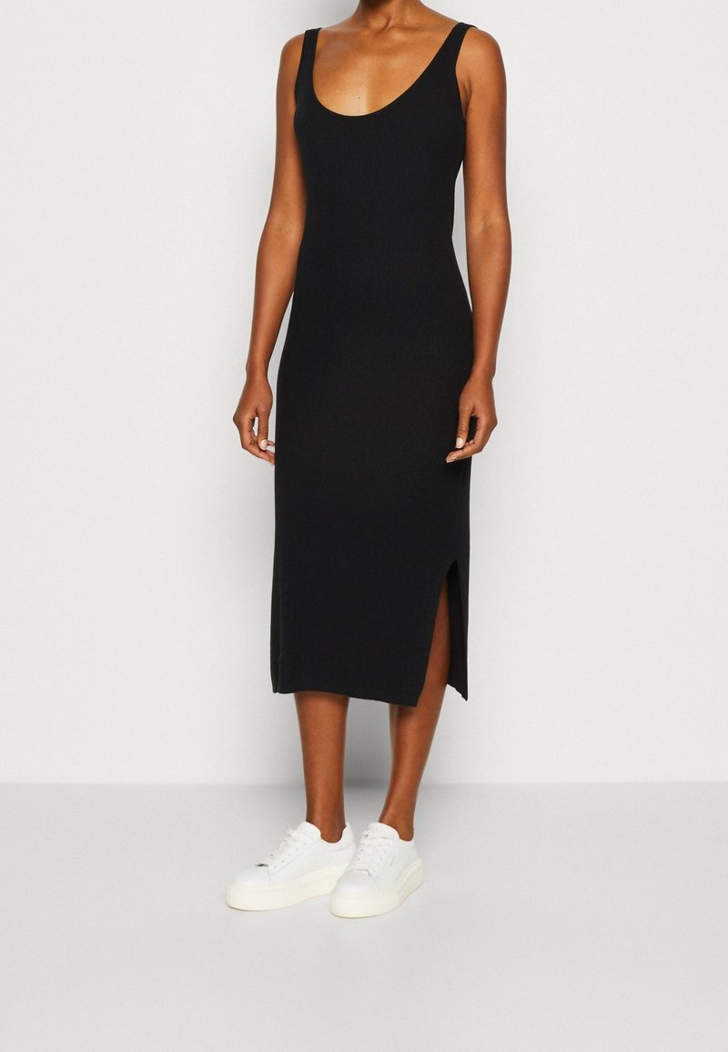 Femme portant une robe midi noire sans manches avec une fente latérale, associée à des baskets blanches, debout devant un fond clair uni.