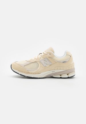 Zapatilla deportiva beige con parte superior de ante y malla, acentos blancos, suela texturizada y un prominente logo N en el lateral. Presenta un collar acolchado.