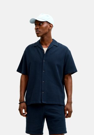 Hombre con camisa azul marino de manga corta y pantalones cortos a juego, gorra azul claro, de pie contra un fondo liso.