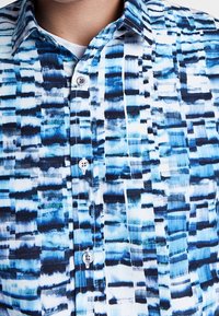 Camicia a maniche corte con bottoni, caratterizzata da un motivo abstract blu e bianco, realizzata in tessuto leggero con colletto regolare e bottoni coordinati.