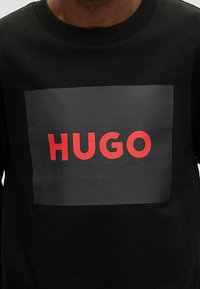 Sudadera negra con un gran logo rectangular en gris oscuro con la palabra "HUGO" en letras rojas y negrita centradas en la parte delantera.