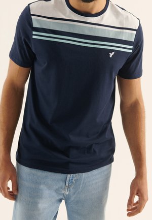 Homme portant un t-shirt bleu marine avec des rayures horizontales blanches et bleu clair sur la poitrine, associé à un jean bleu clair.