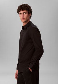 Čierna košeľa na zapínanie s golierom, dlhými rukávmi a úzkym strihom. Hladká látka s jemným leskom bez viditeľného vzoru.