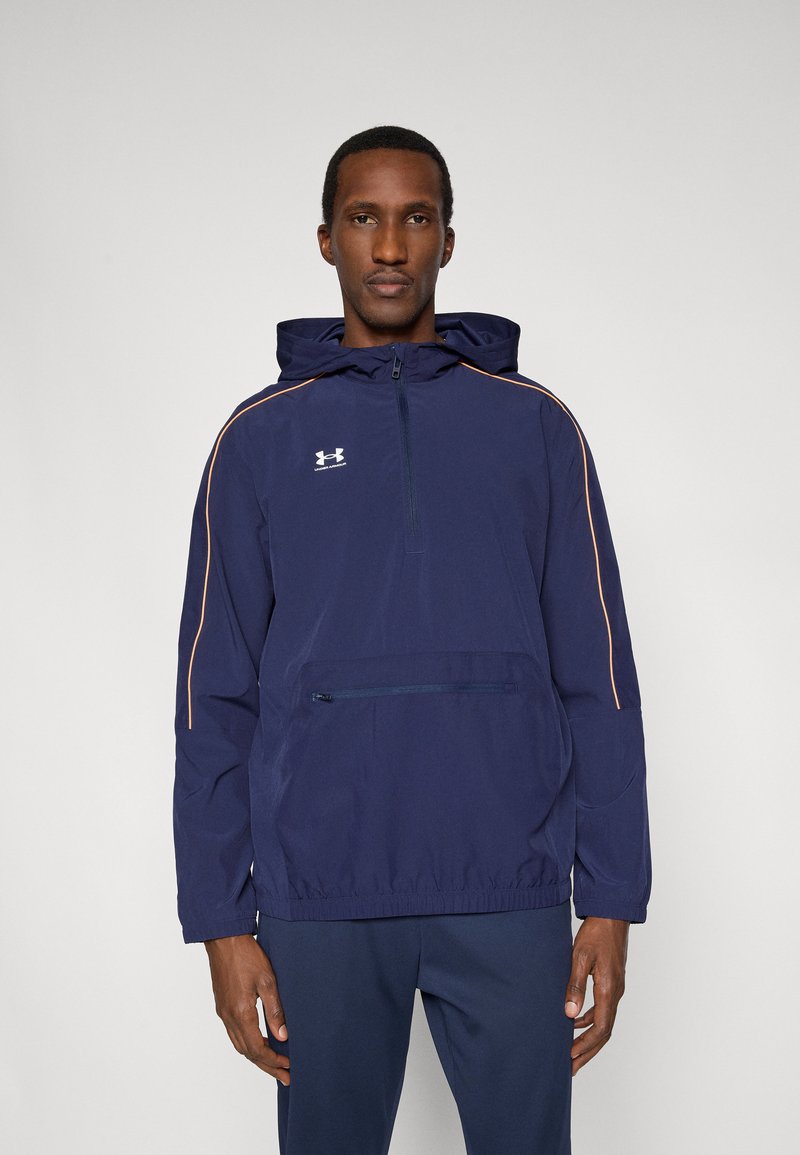 Άνδρας που φοράει μπλε σκούρο μπουφάν Under Armour με πορτοκαλί ραφές και αντίστοιχο μπλε σκούρο παντελόνι, στέκεται μπροστά σε απλό φόντο.