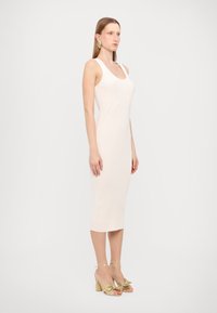Mikroskohtäbeane midi-kleid light beige färgu, med scoop-kragen och diskreta sömmar, kombinerat med guld blockklack-sandaler och rosett-accents.