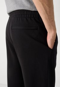 Sort sweatpants med struktureret stof, elastisk talje og en enkelt baglomme. Side sømdetaljer synlige.