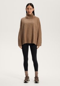 Maglione a collo alto marrone, oversize, con colletto e orlo a coste, abbinato a leggings neri e sneakers marroni con dettagli in tessuto testurizzato.