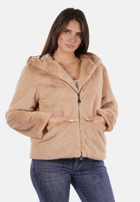Giacca di pelliccia sintetica beige con cappuccio, chiusura con zip e tasche frontali. Ha una vestibilità comoda e una texture morbida. Indossata con jeans casual.