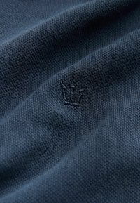 Tissu texturé bleu foncé avec un motif tissé, comportant un petit logo de couronne brodé dans une teinte légèrement plus claire.