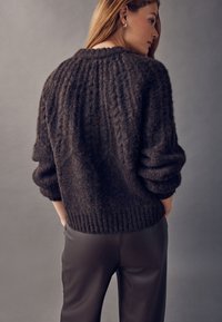 Mørkebrun kabelstrikket sweater med tekstureret finish og ribbet kant, kombineret med slanke sorte bukser, set bagfra.