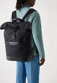 Mochila negra tipo roll-top con correas ajustables, bolsillo lateral y detalles en cuero. Tela de superficie texturizada para mayor durabilidad.