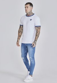 Fehér, rövid ujjú póló kék részletekkel az ujjakon és a nyakvonalon, világoskék, slim-fit farmerrel, kopott részletekkel, és fehér sportcipővel párosítva.