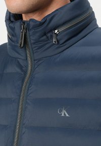 Navy blue padded jacket with a high collar, featuring a central zipper and horizontal stitching. Contains a subtle logo on the chest.
