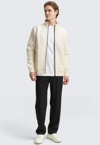 Lichtcrème zip-up jack met een textuurpatroon, zwarte rits, gedragen over een wit T-shirt, gecombineerd met zwarte broek en witte sneakers.