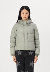 Veste rembourrée verte avec capuche, conçue avec une fermeture éclair à l'avant et un cordon de serrage. Portée avec un jean noir à motifs étoilés.