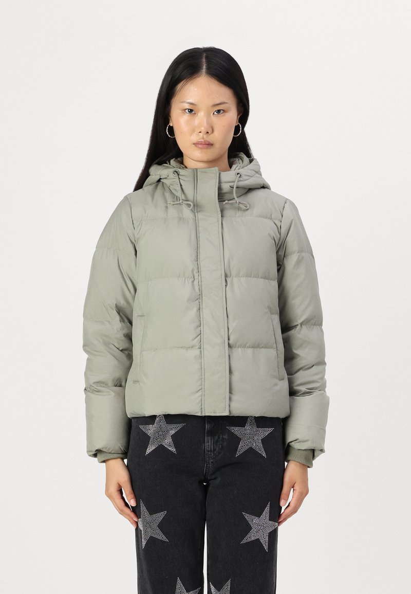 Veste rembourrée verte avec capuche, conçue avec une fermeture éclair à l'avant et un cordon de serrage. Portée avec un jean noir à motifs étoilés.