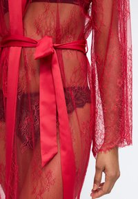 Κόκκινο διάφανο δαντελένιο ρόμπα δεμένη με σατέν ζώνη πάνω από μαύρα δαντελένια εσώρουχα, εμφανίζοντας τον κορμό και το δεξί χέρι με φυσικό τόνο δέρματος.