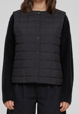 Kvinde iført sort quiltet vest med trykknapper over en sort langærmet sweater og sorte bukser, vist fra skuldre til hofter.
