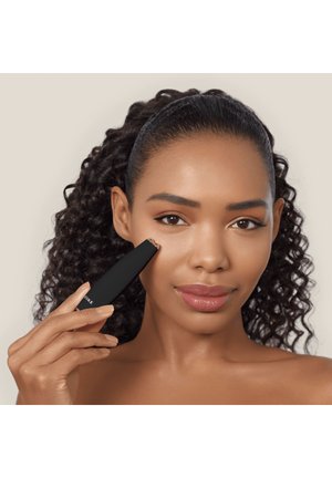 Femme aux cheveux bouclés utilise un épilateur facial noir près de la joue, montrant une peau lisse et un maquillage naturel.