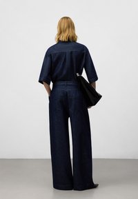 Outfit in denim caratterizzato da una maglietta ampia e pantaloni a gamba larga in azzurro scuro. Accentuato con una borsa nera, visto da dietro.