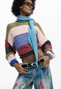 Jersey de punto colorido con varias texturas y rayas horizontales, combinado con vaqueros multicolores salpicados de pintura y un cinturón negro.