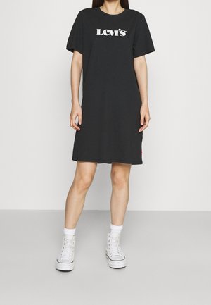 Mujer vistiendo un vestido camiseta negro de Levi's con logo blanco, calcetines blancos y zapatillas blancas de caña alta, de pie contra un fondo liso.