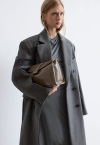 Grau gestrickter Mantel mit übergroßen Revers, getragen über einem grauen, drapierten Kleid; hält eine glänzende, rechteckige braune Clutch.