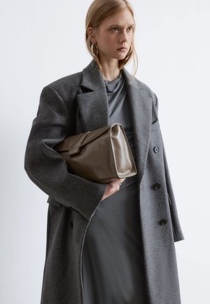 Grau gestrickter Mantel mit übergroßen Revers, getragen über einem grauen, drapierten Kleid; hält eine glänzende, rechteckige braune Clutch.