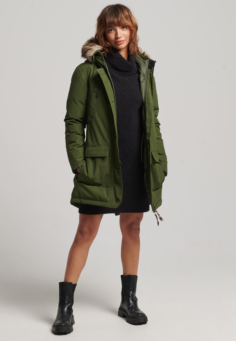 Frau in einer olivgrünen Parka mit Fellkapuze, schwarzem Strickkleid und schwarzen Knöchelstiefeln, die vor einem schlichten grauen Hintergrund steht.