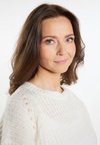 Biała dziergana sweter z teksturowanym wzorem i okrągłym dekoltem, z ażurowymi akcentami na ramionach, noszona przez kobietę z falującymi włosami.