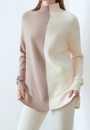 Mujer con un suéter de cuello alto con mitades verticales en beige y crema, combinado con pantalones de color claro, manos sosteniendo el dobladillo del suéter.