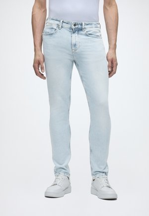 Άνδρας με ανοιχτό μπλε skinny τζιν και λευκά αθλητικά παπούτσια, στέκεται μπροστά σε απλό λευκό φόντο.
