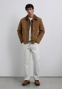 Chaqueta de ante marrón con cierres tipo toggle y un cuello marrón, combinada con pantalones blancos y botas de ante marrón, de pie contra un fondo neutro.