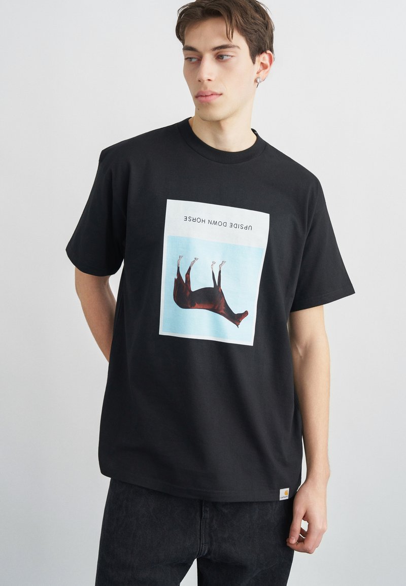 Νεαρός άνδρας που φοράει μαύρο T-shirt με γραφικό ενός αναποδογυρισμένου αλόγου και το κείμενο «UPSIDE DOWN HORSE», στέκεται μπροστά σε απλό φωτεινό φόντο.