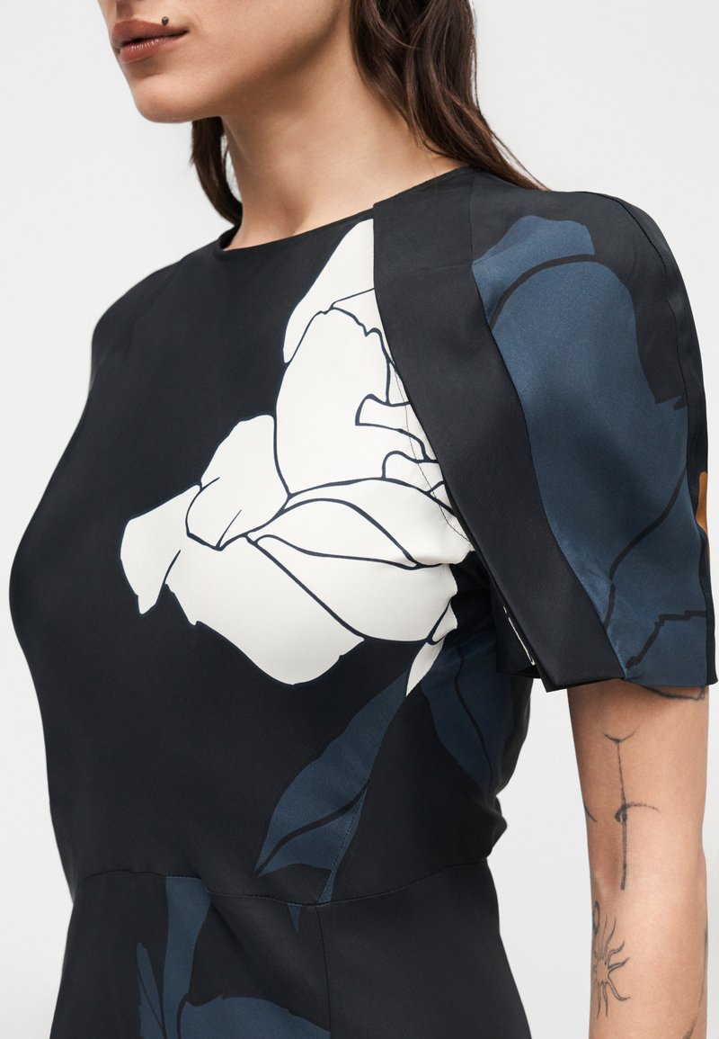 Femme portant une robe noire avec un imprimé floral blanc et bleu large, manches courtes bouffantes, et tatouages visibles sur les bras.