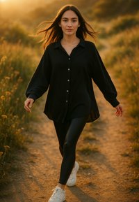 Camicia nera con bottoni, maniche larghe e orlo asimmetrico, abbinata a leggings neri aderenti e sneakers bianche, mentre cammina su un sentiero di terra.