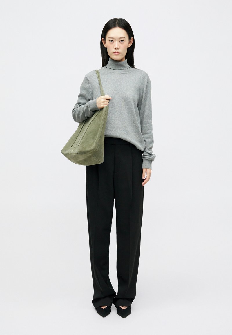 Pull à col roulé gris, pantalon large noir et sac à bandoulière en daim vert. Le modèle se tient droit, portant des chaussures à talons noires.
