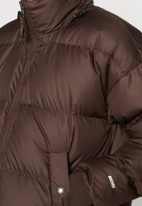 Braune Puffjacke mit hohem Kragen, glänzender Oberfläche, Reißverschluss und elastischen Bündchen. Verfügt über sichtbare Nähte und ein Logo am Ärmel.