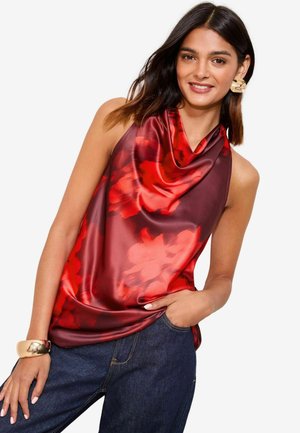 Haut sans manches rouge et bordeaux en satin, avec un décolleté en cowl fluide et un motif floral, associé à un jean en denim foncé.
