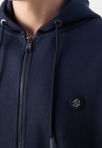 Gros plan sur une personne portant un sweat à capuche zippé bleu marine avec une capuche et un logo circulaire noir sur la poitrine.