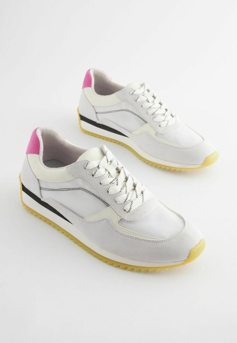 Ein Paar weiße und beige Sneakers mit pinken Fersenakzenten, schwarzen Streifen, weißen Schnürsenkeln und gelben strukturierten Sohlen auf einem weißen Hintergrund.