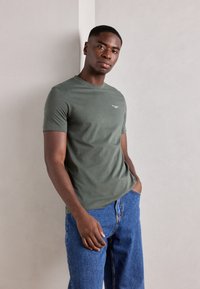 Tricou din bumbac de culoare verde măsliniu, cu mâneci scurte, guler rotund și un branding mic alb pe piept, purtat cu pantaloni din denim albastru deschis.