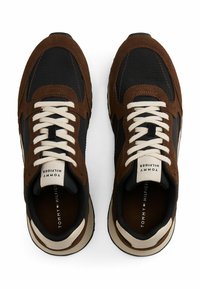 Zapatillas marrones y negras con materiales de ante y textil, que cuentan con cordones crema, una etiqueta con el logo en la lengüeta y una suela de goma texturizada.